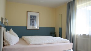 Ein ordentlich bezogenes Bett mit Kissen, ein gerahmtes Bild an der Wand und ein Fenster mit Vorhängen.