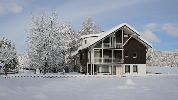 Eine zweistöckige Chalet mit Balkon, umgeben von schneebedeckten Bäumen und blauem Himmel.