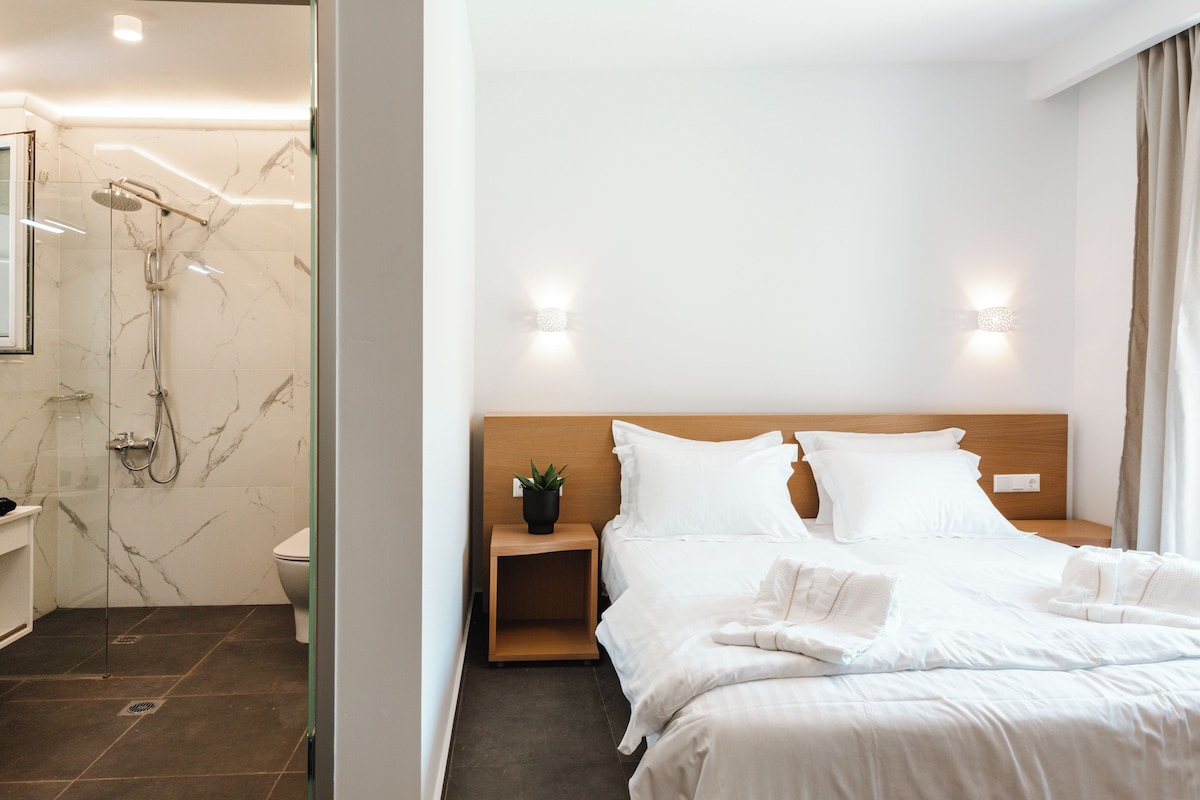 A hotel room with a marble shower, a wooden headboard, white bedding, and a small bedside table with a plant.
