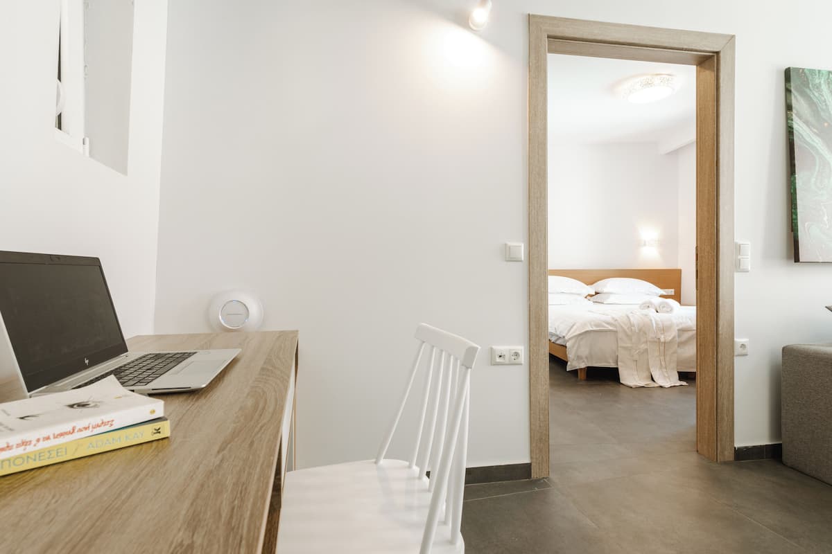 A room with a wooden desk, a laptop, and books. There is a bed with white bedding and a wooden headboard.
