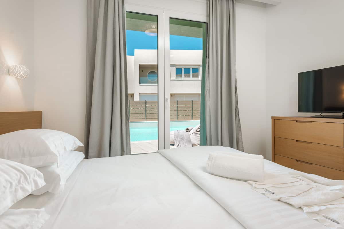 A hotel room with a bed, a TV, a wooden dresser, and a view of a pool and building through a glass door.