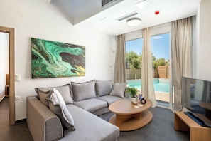 Un salon moderne avec un canapé d’angle, une table basse ronde, un téléviseur à écran plat et une grande fenêtre donnant sur une piscine.