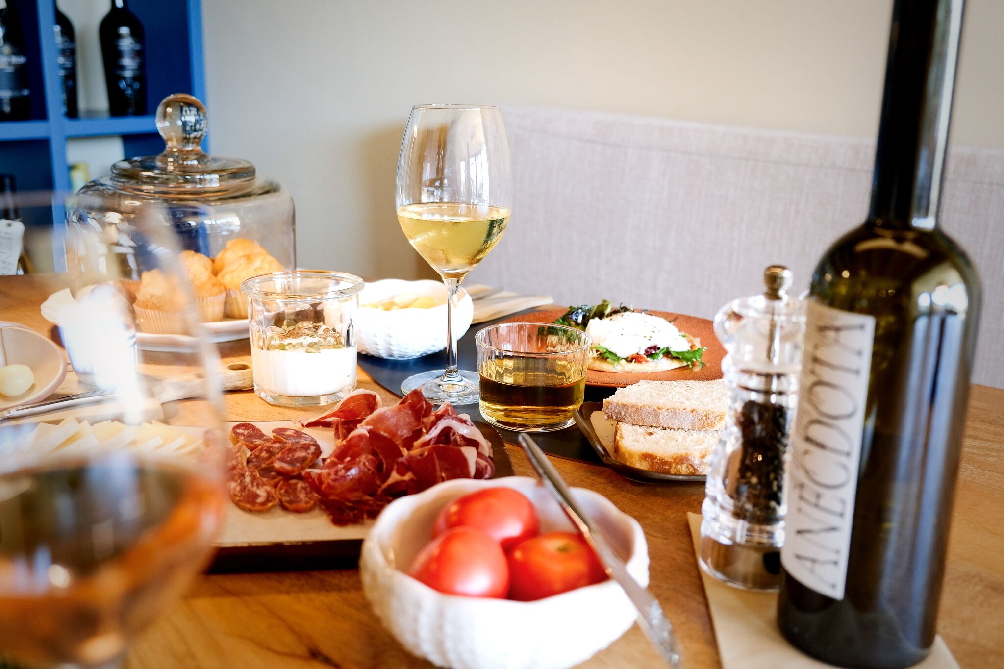 A table with various food items, including bread, cheese, and tomatoes, along with glasses of wine and a bottle of olive oil.