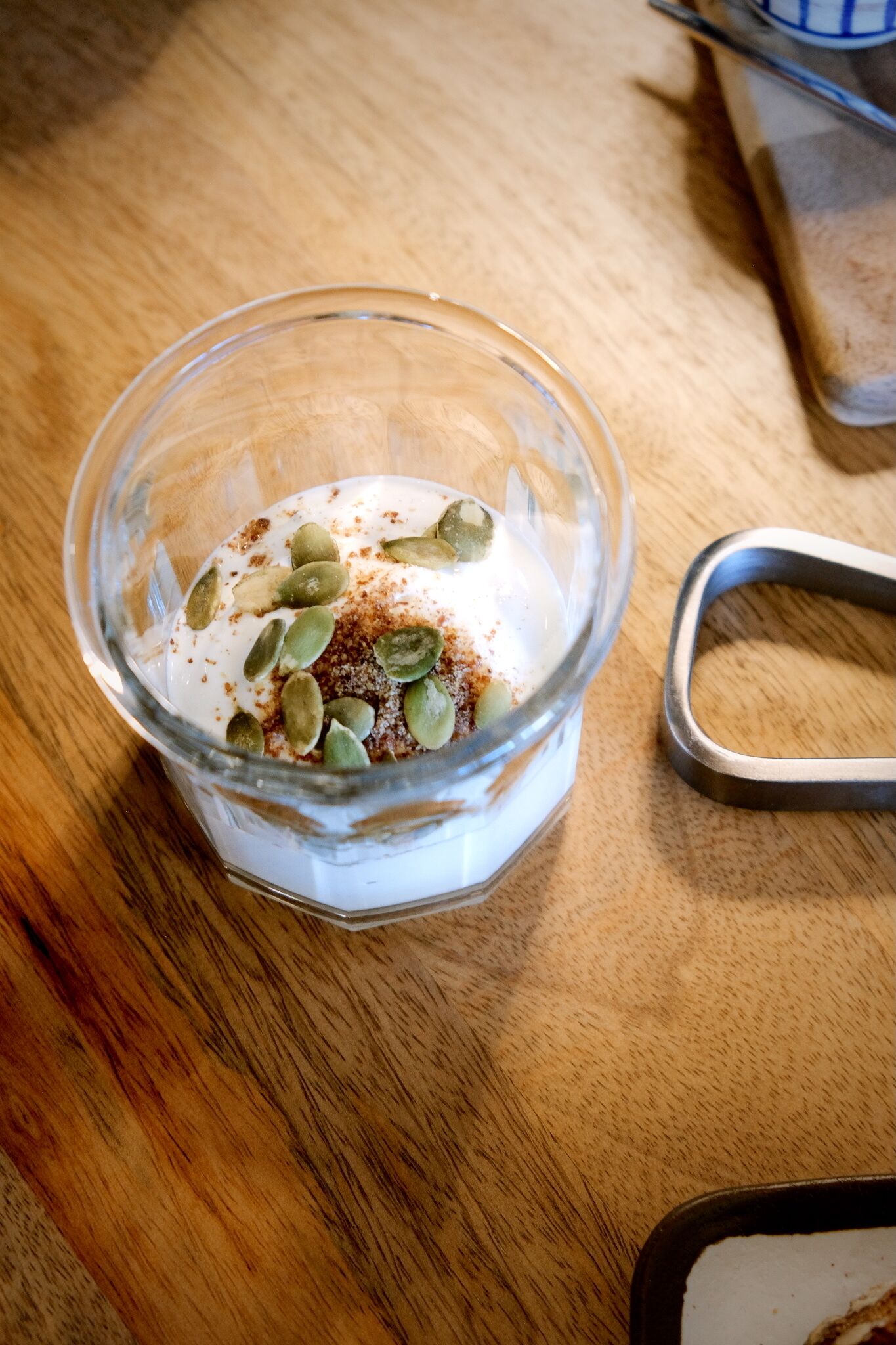 A glass bowl containing a creamy substance with pumpkin seeds and a brown spice on a wooden surface.