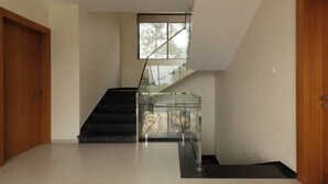 A modern interior with a glass staircase leading to a landing, a wooden door on the left, and a plain wall on the right. - Brick and beam (Mahabaleshwar)