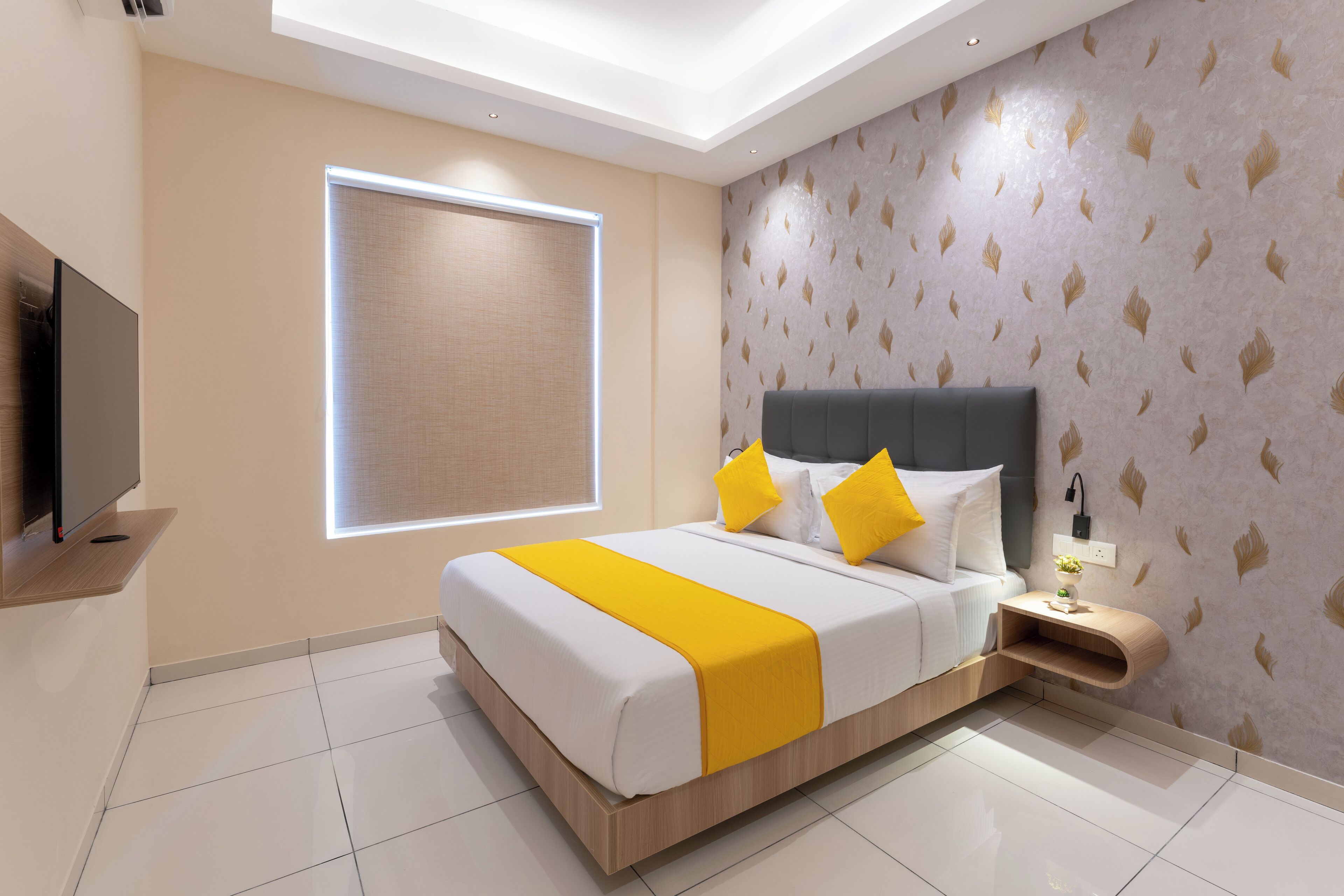 A modern hotel room with a large bed, a flat-screen TV, a bedside table with a lamp, and a window with blinds.