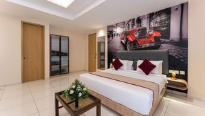 A modern hotel room with a large bed, a small side table with a vase of flowers, a wall-mounted TV, and a cityscape mural. - Hotel Rivido Near Manyata Tech park (Bengaluru)
