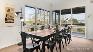 Un espace repas avec une longue table et des chaises, de grandes fenêtres et une affiche encadrée au mur.