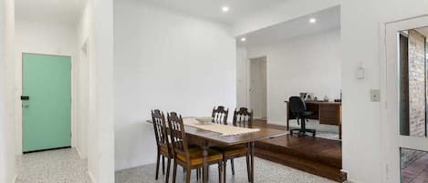 A spacious room with a dining table and chairs, a desk with a chair, and a green door.
