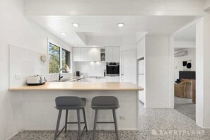Une cuisine moderne avec des meubles blancs, un grand îlot central doté de deux tabourets de bar et une vue sur l’extérieur par la fenêtre.