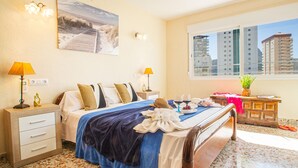 A hotel room with a bed, bedside tables, a window with a view of buildings, and a beach scene on the wall. - Villa Catrina - Plusholidays (Calpe)