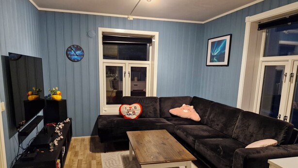 Una sala de estar con un sofá negro, una mesa de centro de madera, un televisor sobre un mueble, un reloj y una ventana con vistas al exterior.