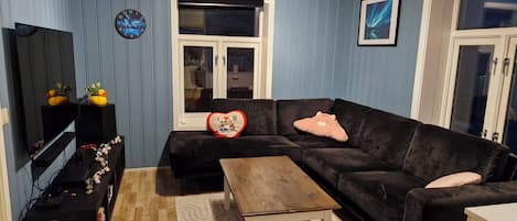 Sala de estar com sofá preto, mesa de centro de madeira, televisão em um suporte, relógio e janela com vista para o exterior.