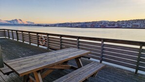 Una terraza de madera con una mesa de picnic y un flotador salvavidas, con vistas a un río y un paisaje urbano al fondo.