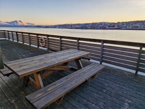 Eine Holzterrasse mit Picknicktisch und Rettungsboje, mit Blick auf einen Fluss und eine Stadtkulisse im Hintergrund.