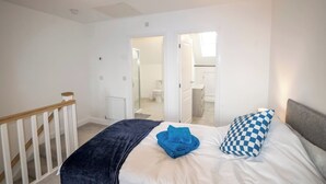 A bedroom with a bed, a blue towel, and a checkered pillow. There is a white radiator and a wooden staircase. - A stunning new build property (Cheshire West and Chester)