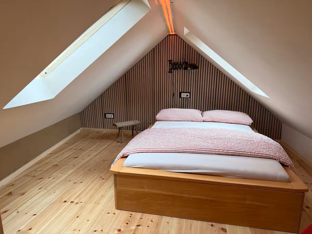 Ein Schlafzimmer mit schräg stehender Decke, Holzboden, einem Bett mit gestreifter Bettwäsche und einem kleinen Tisch.