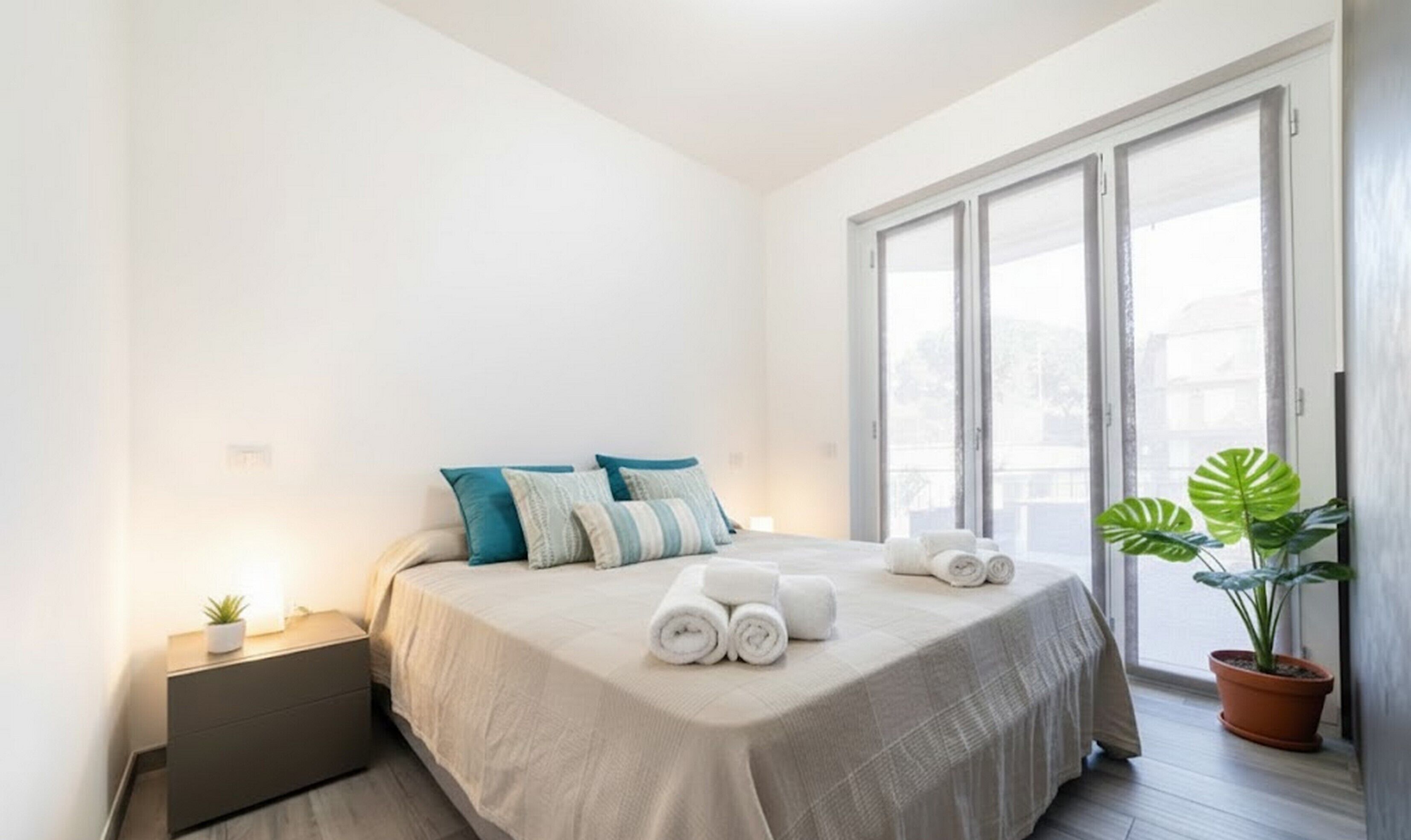 A bedroom with a bed, pillows, a nightstand, a potted plant, and a window with curtains.