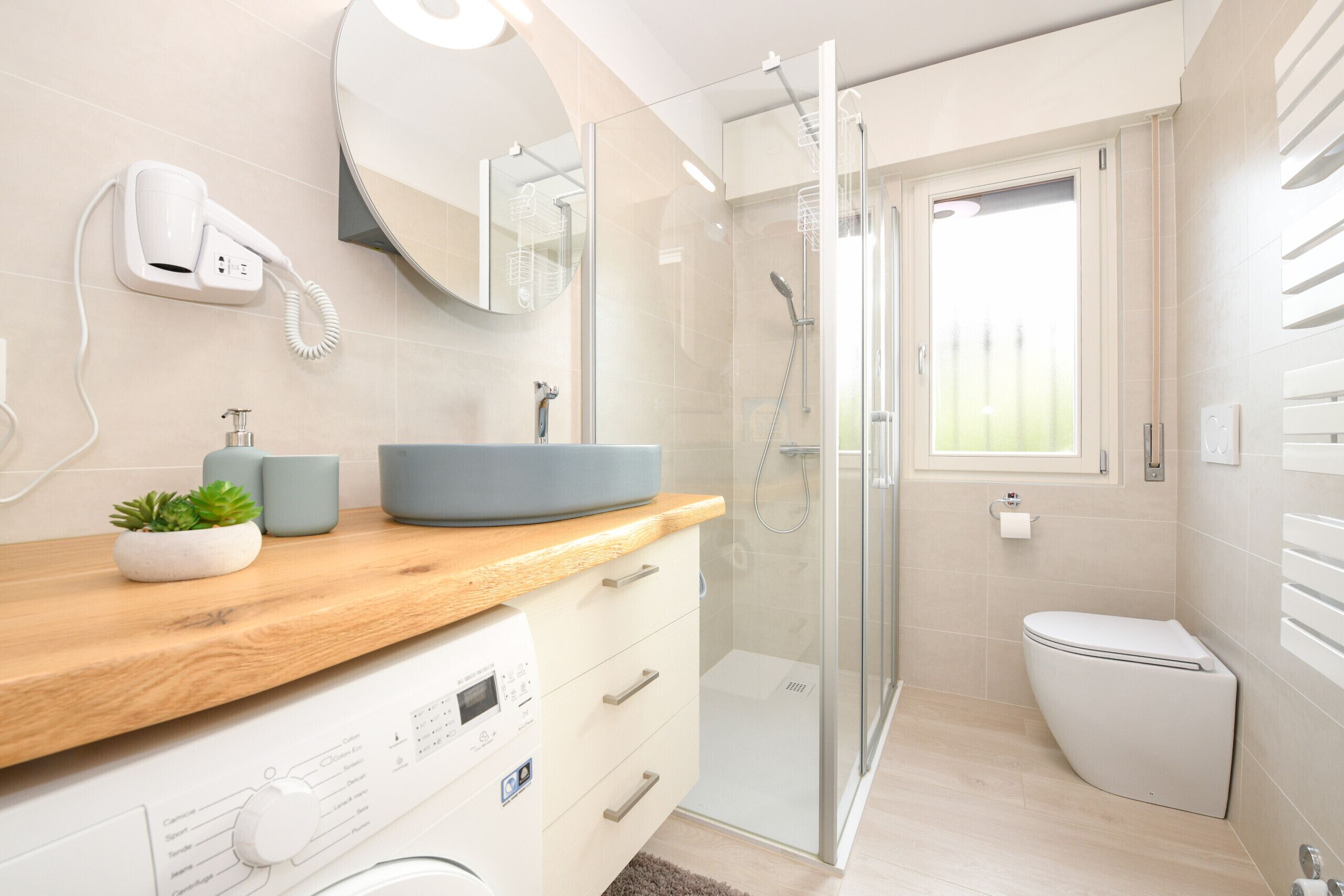 A modern bathroom with a glass shower enclosure, a sink with a blue basin, a hairdryer mounted on the wall, and a toilet.