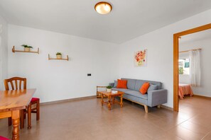 A living room with a wooden dining table, a grey sofa with orange cushions, a wooden coffee table, and a ceiling light fixture. - Villa Mercedes - Plusholidays (Calpe)