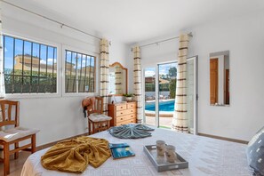 A bedroom with a bed, a chair, a dresser, a window, and a view of a swimming pool. - Villa Mercedes - Plusholidays (Calpe)