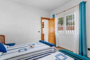 A bedroom with two beds, a wooden door, a window with a view of a balcony, and a chair. - Villa Mercedes - Plusholidays (Calpe)
