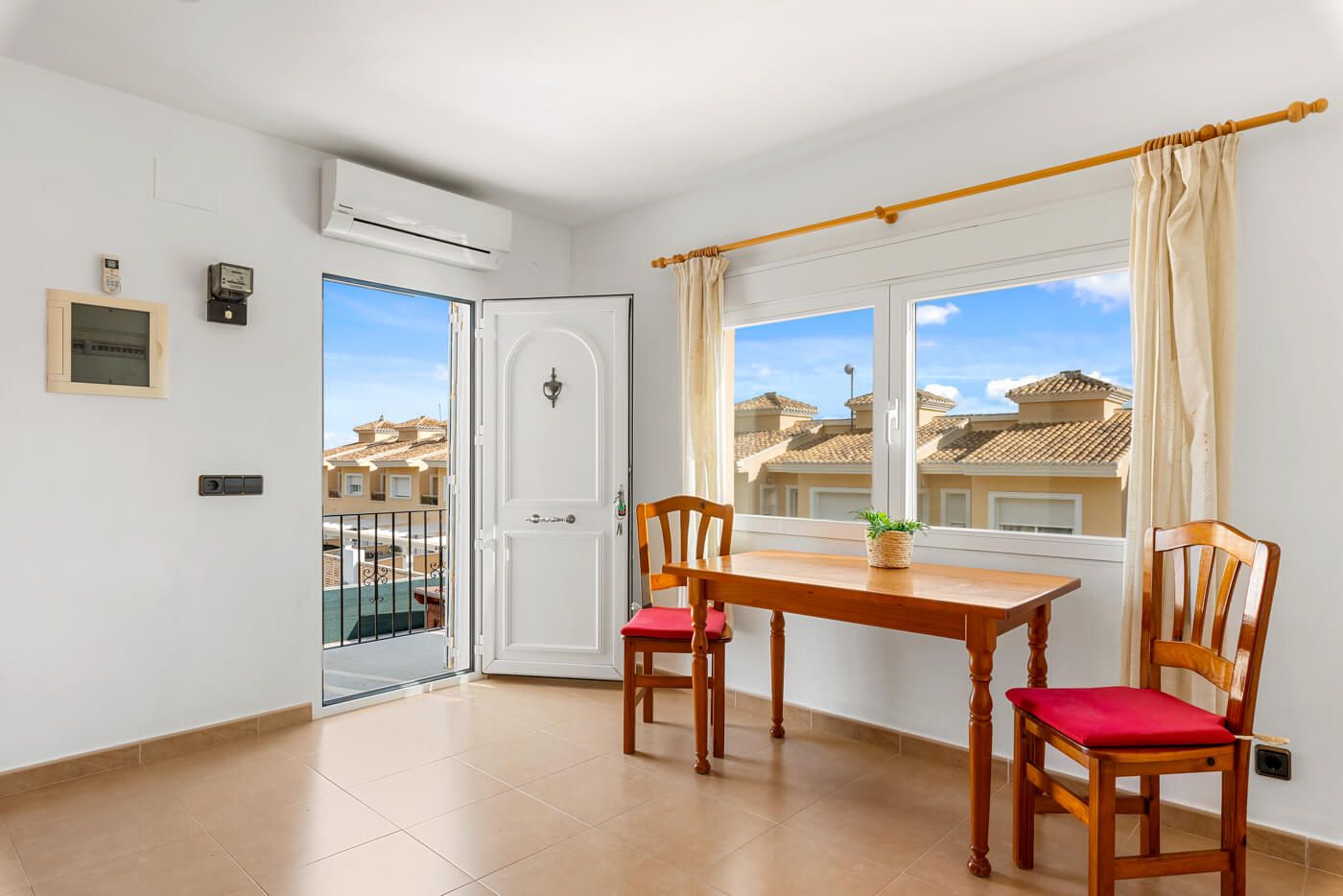 A room with a wooden table and chairs, a window with curtains, an air conditioning unit, and a door leading outside.