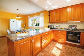 A kitchen with wooden cabinets, a granite countertop, a refrigerator, a microwave, and an oven. - Villa Flora - Plusholidays (Calpe)