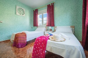 A room with two beds, a window with curtains, a wall clock, and a small table with a lamp. - Villa Flora - Plusholidays (Calpe)