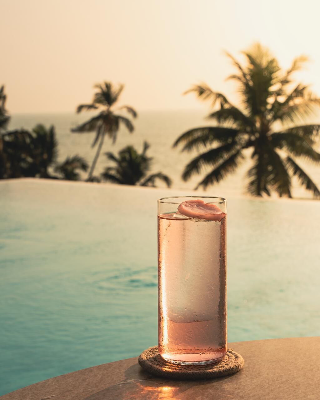 A glass of a clear liquid with a slice of fruit on a coaster by a pool with palm trees in the background.