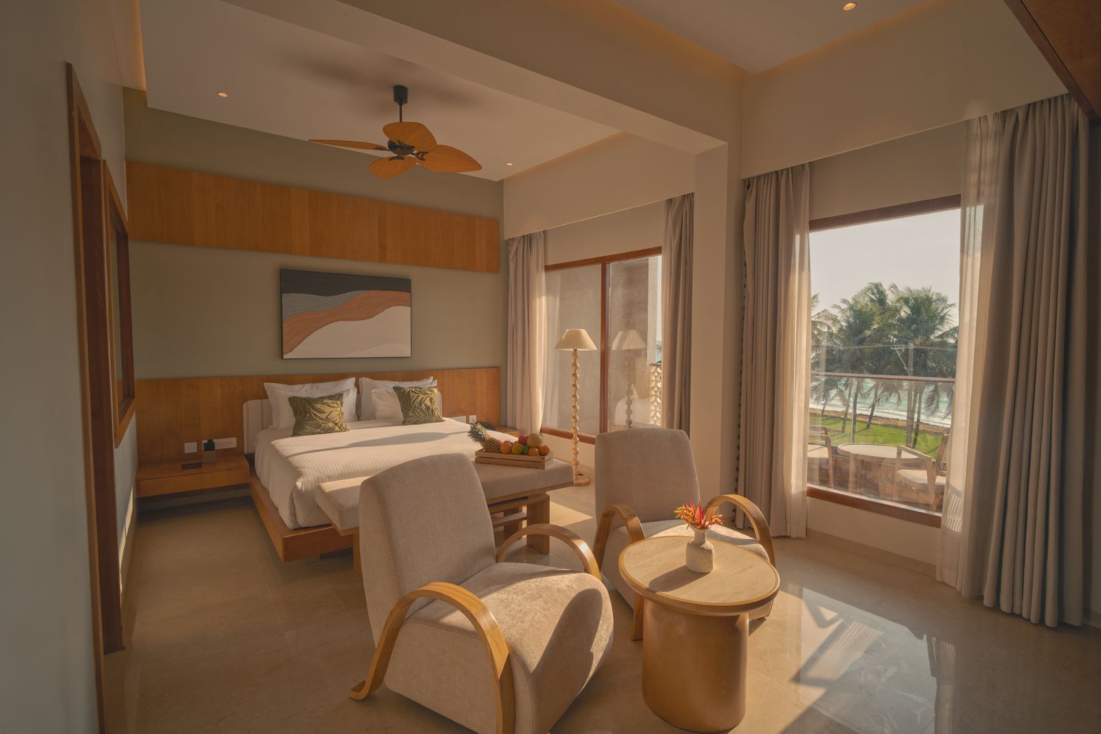 Une chambre d’hôtel moderne équipée d’un lit, de deux fauteuils, d’une petite table, d’un ventilateur de plafond et d’une grande fenêtre donnant sur des palmiers.
