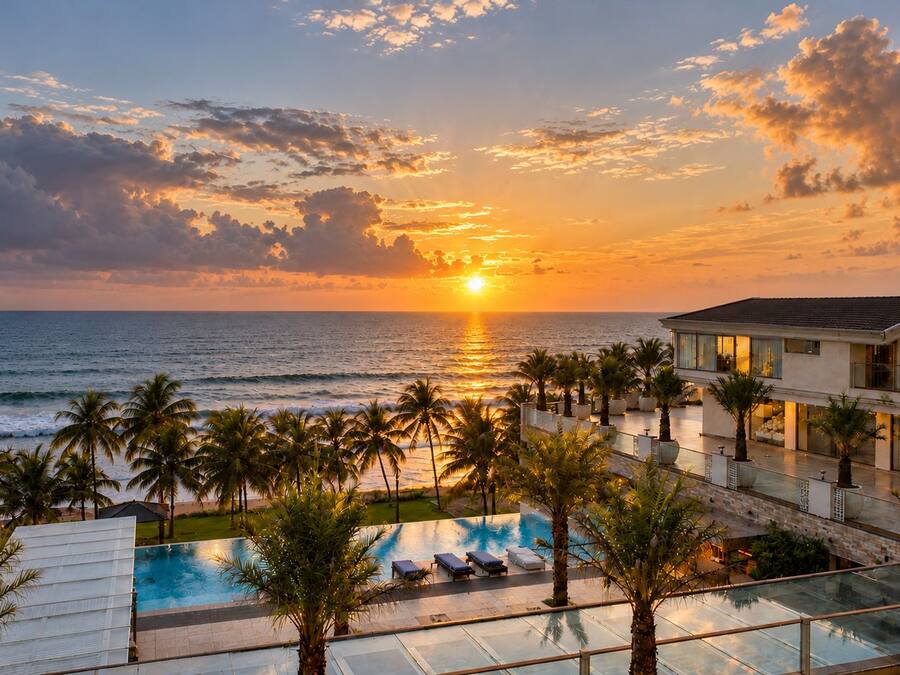 Un resort con piscina, palmeras y vista al océano al atardecer.