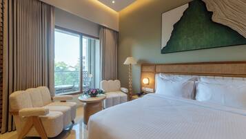 Habitación de hotel con una cama grande, dos sillones, una mesita con un jarrón de flores, una mesita de noche con lámpara y vistas al exterior a través de un ventanal.