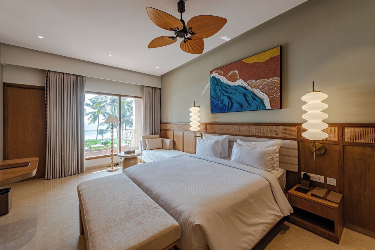 A hotel room with a large bed, a ceiling fan, a painting, and a view of palm trees.