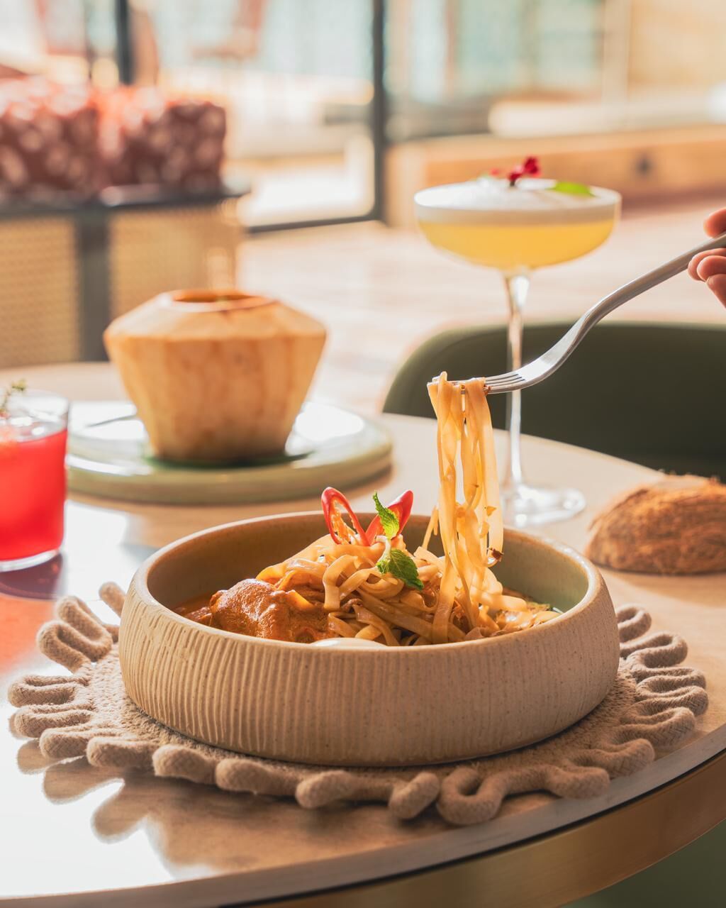 A bowl of pasta with a fork lifting some noodles, a glass of a yellow drink, and a pastry on a table.