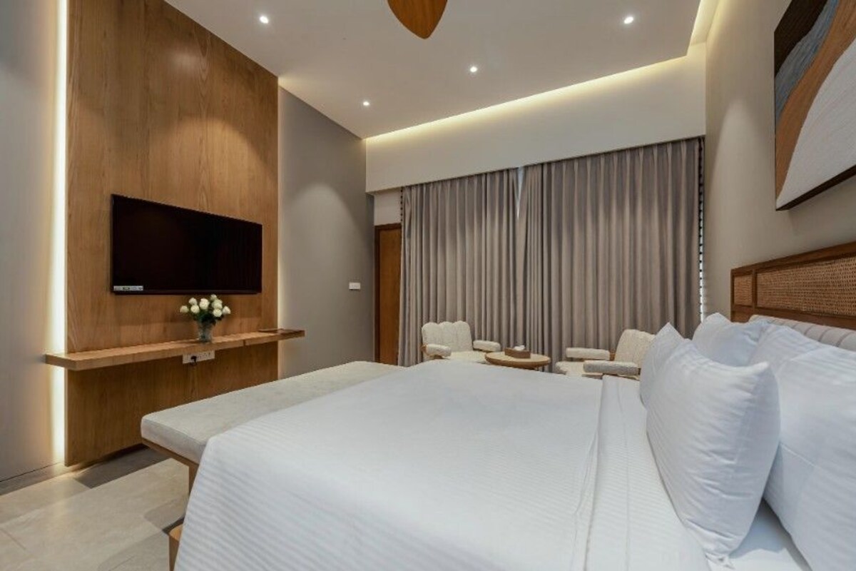 A modern hotel room with a large bed, a flat-screen TV mounted on a wooden panel, a seating area with chairs and a small table, and a large window with curtains.