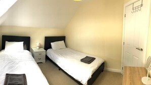 A small, well-lit bedroom with two single beds, a bedside table with a lamp, and a door with a coat rack. -  Cosy warm cottage Cirencester,Capital of the Cotswolds,bibury close pet frendly (Cirencester)