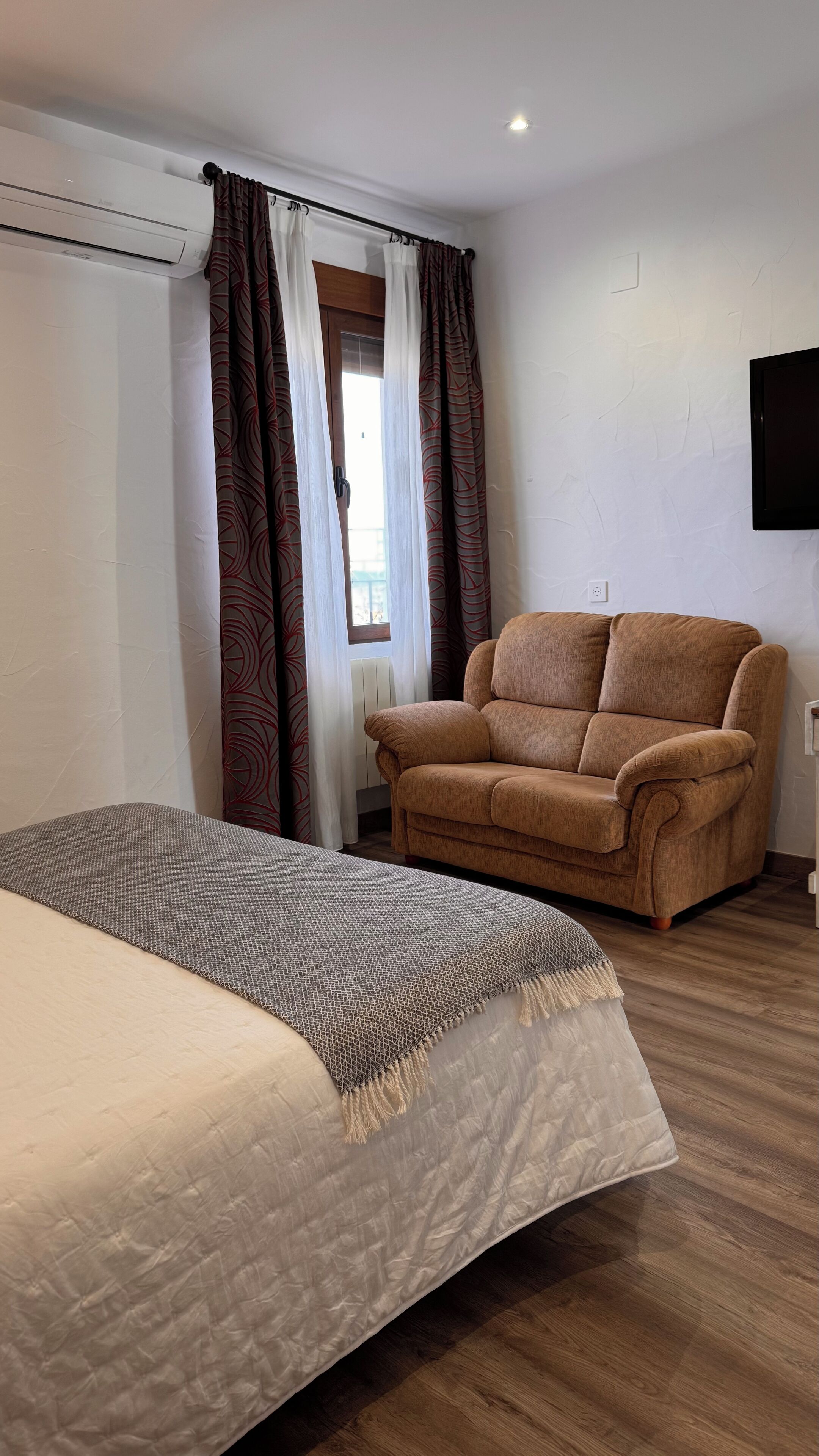Una habitación con una cama, un sillón marrón y una ventana con cortinas estampadas.