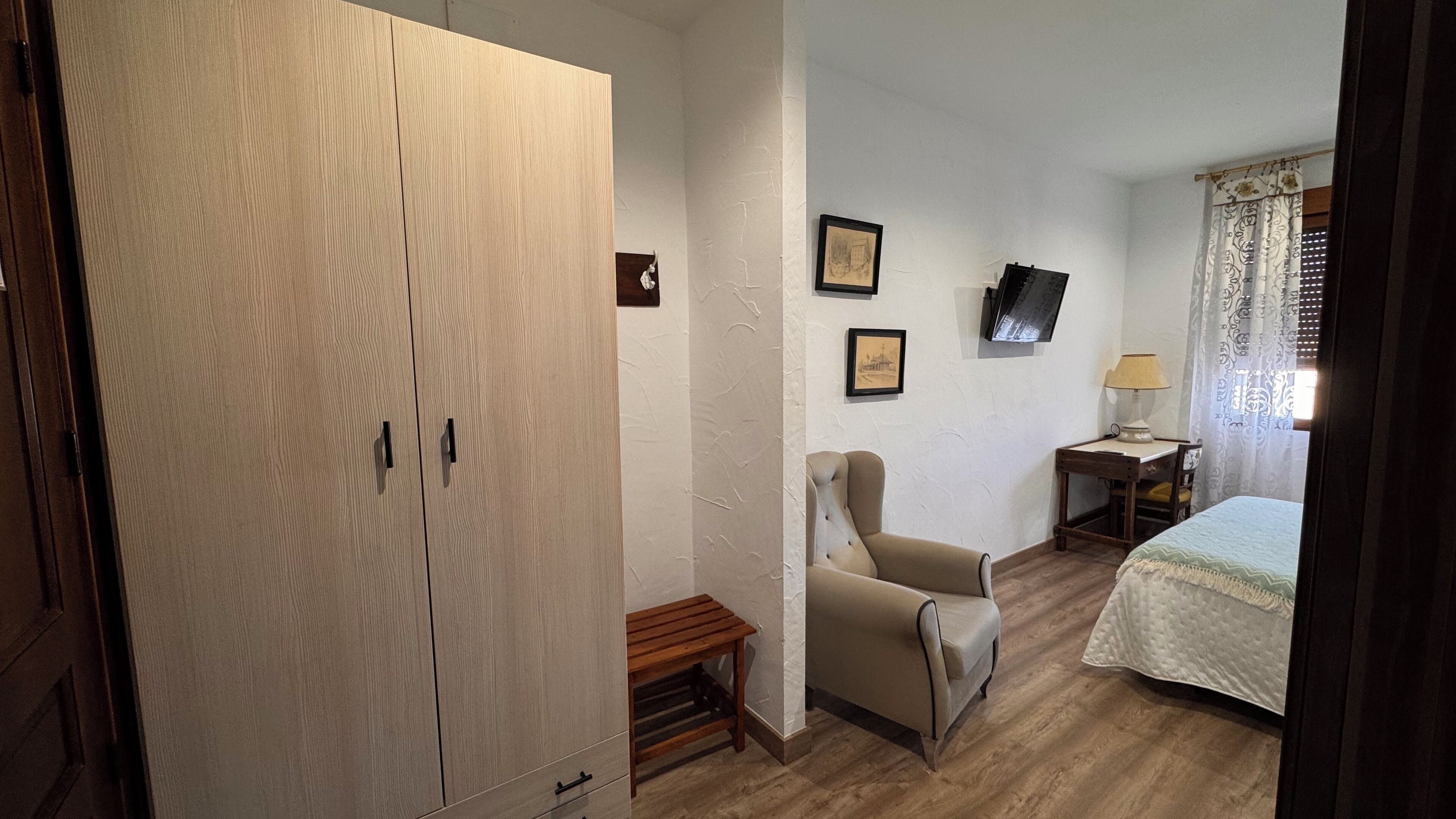 Quarto com guarda-roupa de madeira, cama com colcha, cadeira, mesa pequena com abajur e duas imagens emolduradas na parede.