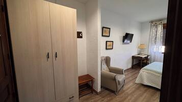 Quarto com guarda-roupa de madeira, cama com colcha, cadeira, mesa pequena com abajur e duas imagens emolduradas na parede.