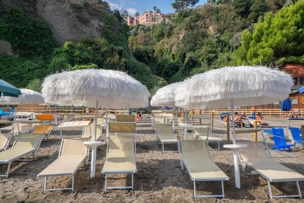 Une plage avec des transats et des parasols en chaume, une colline rocheuse en arrière-plan, et quelques personnes au loin.