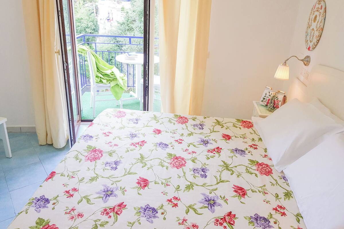 Une chambre à coucher avec une couvre-lit à motifs floraux, une vue sur un balcon avec une chaise et une table, et une table de chevet avec une lampe et un objet décoratif.