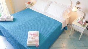 Un lit bien fait, recouvert d’une couette bleue, avec des oreillers blancs et des serviettes ornées de rubans roses. Il y a deux tables de chevet avec des lampes et une assiette décorative accrochée au mur.
