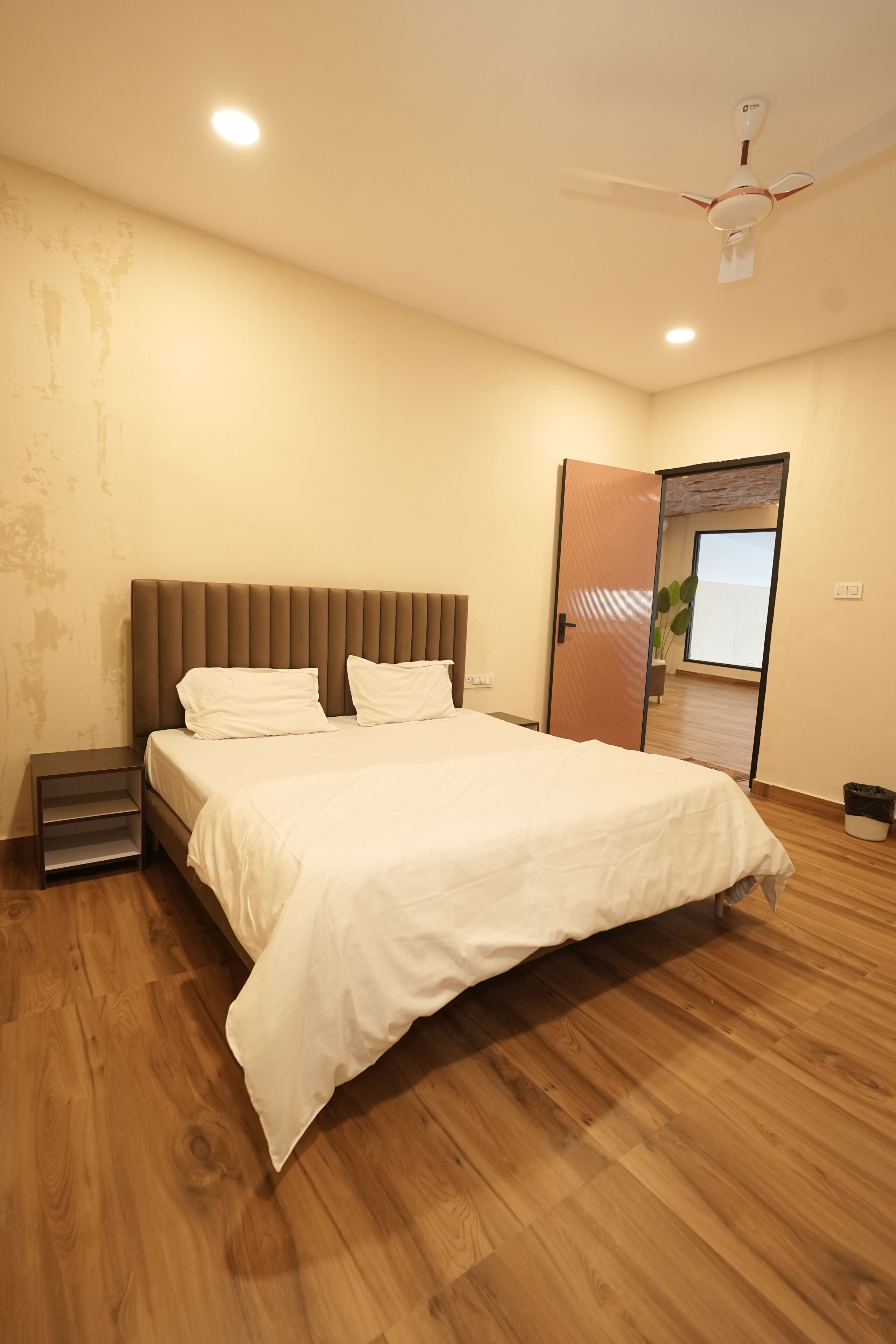 A bedroom with a bed, wooden flooring, a ceiling fan, and a door leading to another room.