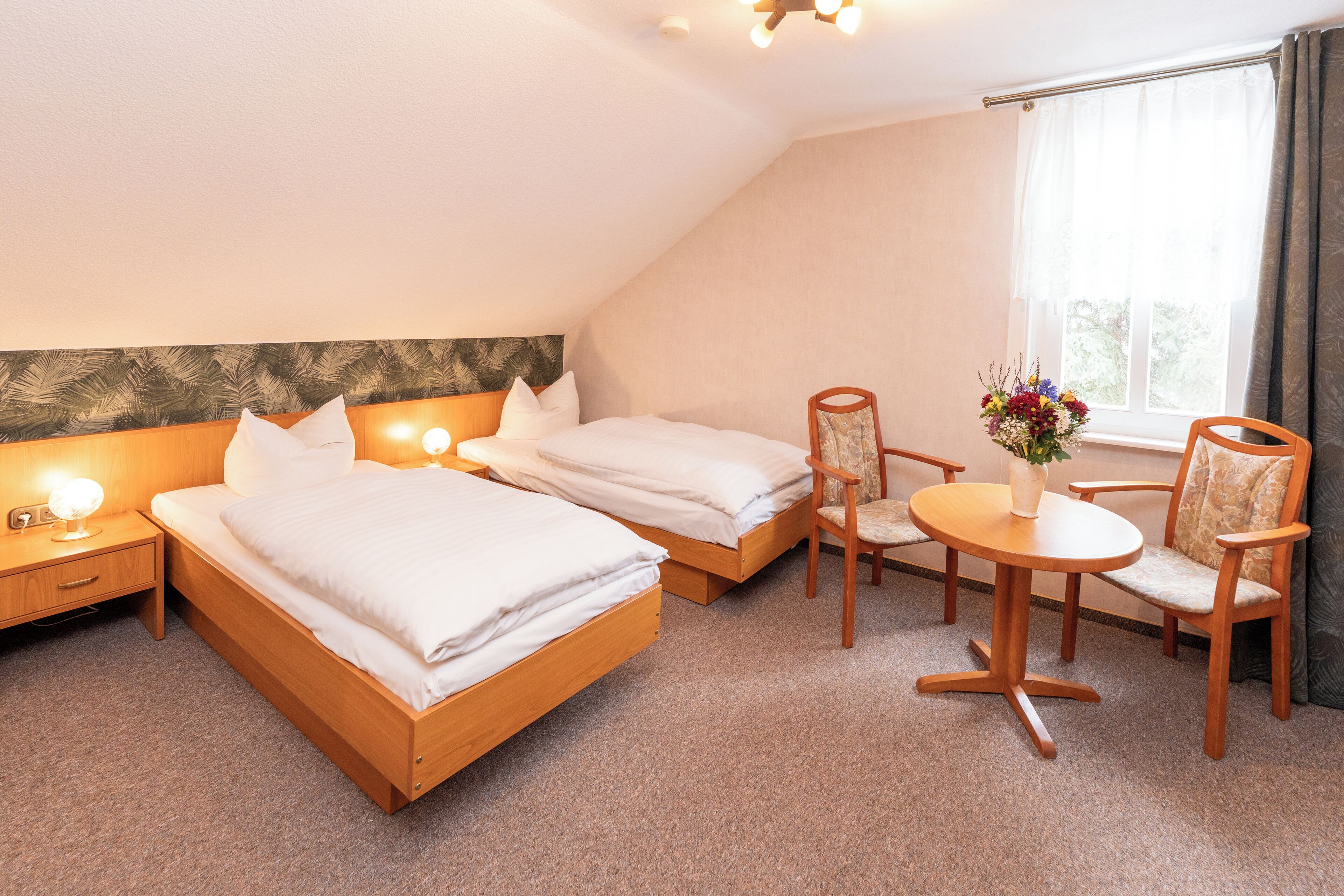 Ein Hotelzimmer mit zwei Betten, einem kleinen Tisch mit Stühlen, einem Nachttisch und einem Fenster mit Vorhängen.