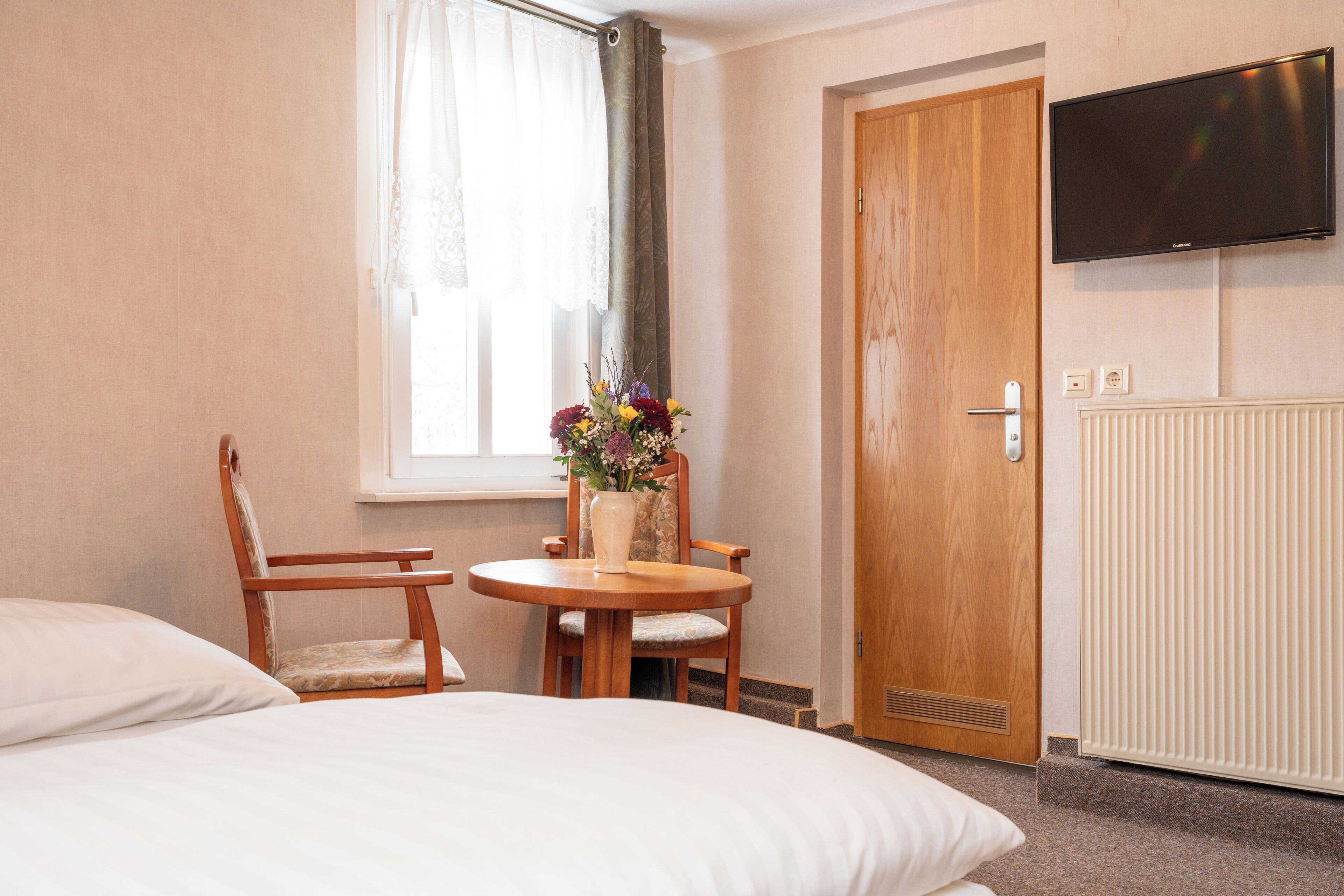 A hotel room with a bed, two wooden chairs, a small table with a vase of flowers, a TV mounted on the wall, and a wooden door.
