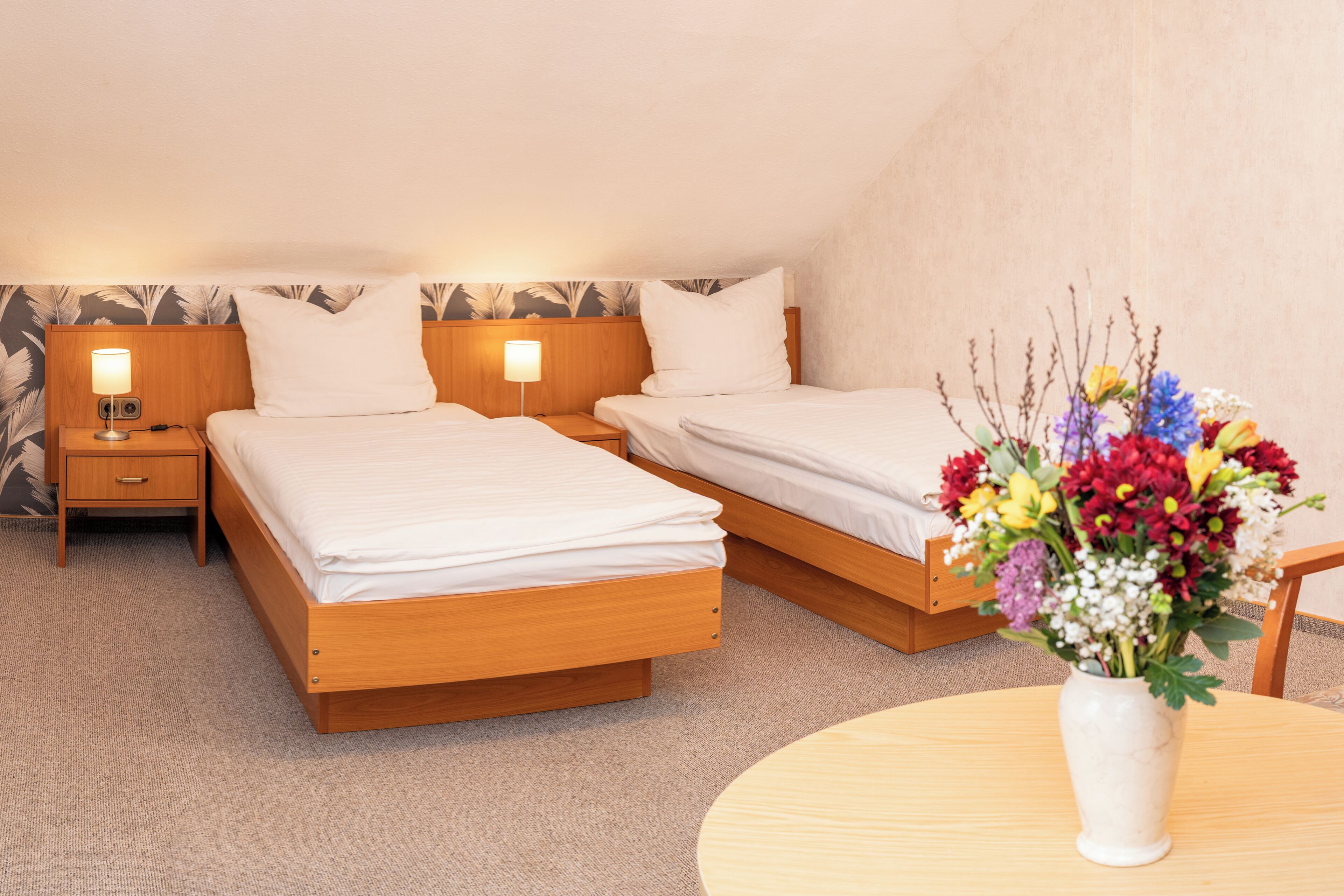 Une chambre d’hôtel avec deux lits simples, une table de chevet avec une lampe, une petite table avec un vase de fleurs et un sol recouvert d’une moquette.