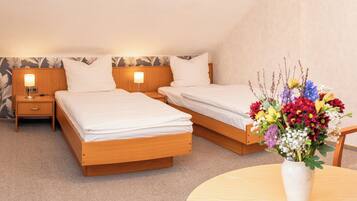 Une chambre d’hôtel avec deux lits simples, une table de chevet avec une lampe, une petite table avec un vase de fleurs et un sol recouvert d’une moquette.