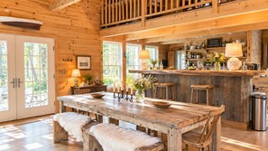 A rustic wooden interior with a dining area, a kitchen with bar stools, and a view of the outdoors through large windows. - French-flair coastal retreat in Port-Clyde (St. George)