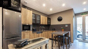 A modern kitchen with a stainless steel refrigerator, wooden cabinets, a built-in microwave, and a coffee machine. There is a small island with stools and a potted plant. - A lovely vacation 5BR/5BATH home with WIFI & AC (L-Imsida)